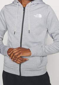 Persona con sudadera de cremallera en gris claro, con cordones blancos y el logo de "The North Face" en el pecho, con las manos entrelazadas al frente.