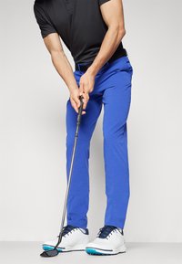 Modré golfové nohavice s prispôsobeným strihom a textúrovanou tkaninou, v kombinácii s čiernym polo tričkom a bielymi golfovými topánkami s modrými akcentmi, v postoji na putting.