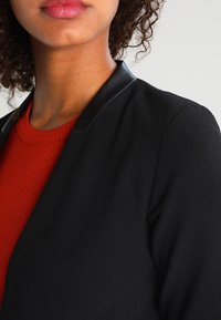 Blazer noir à la texture lisse avec des accents en cuir au col, associé à un haut rouge côtelé. Accent sur des lignes nettes et une coupe ajustée.