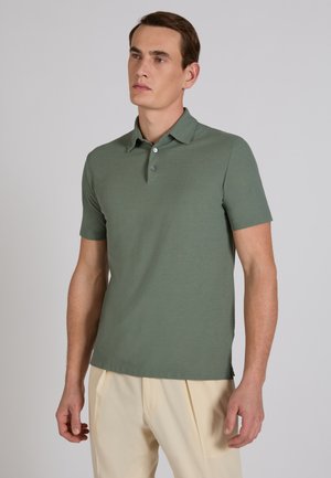 Uomo che indossa una polo a maniche corte verde smorzato e pantaloni beige, in piedi davanti a uno sfondo grigio uniforme.