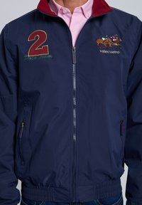 Chaqueta bomber de nailon azul marino con cuello rojo, con detalles bordados: número "2", "Equipo Polo" y jinetes a caballo. Cierre con cremallera.