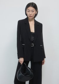 Donna in giacca nera, pantaloni a gamba larga e top di raso che tiene una borsa a mezzaluna nera davanti a uno sfondo bianco semplice.