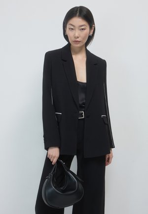 Woman in black blazer, wide-leg pants, and satin top holding black crescent handbag against plain white background.