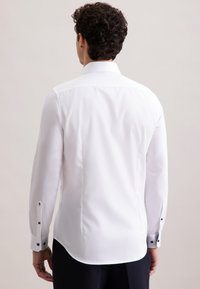 Man met donker krullend haar in een getailleerde witte blouse met lange mouwen en donkere broek, weergegeven vanaf de achterkant tegen een effen achtergrond.