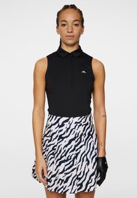 Svart ärmlös polo med krage och logotyp, tillsammans med en vit kjol med marinblå zebra-randningar och veck. Svart handsk på vänster hand.