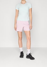 Nainen, jolla on yllään vaaleansininen lyhythihainen paita, vaaleanpunaiset sortsit, valkoiset Puma-sukat ja mustat lenkkarit, seisoo yksiväristä taustaa vasten.