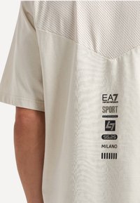 Camicia corta beige chiaro con maniche corte, dotata di una parte inferiore testurizzata e una parte superiore in rete. Presenta loghi e dettagli stampati sui lati.