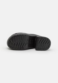 Semelle de chaussure noire avec des motifs de profondeur de bande de roulement et un petit logo embossé au centre, présentée sur un fond clair.