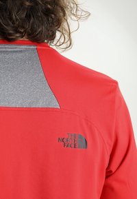 Camiseta atlética roja con un panel de hombro gris, tela texturizada y logo negro en la parte posterior, diseñada para la comodidad y el rendimiento.