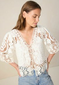 Weiße Spitzenbluse mit V-Ausschnitt, kurzen puffartigen Ärmeln und aufwendigen floralen Mustern. Verfügt über einen Bindebandverschluss und einen gewellten Saum.