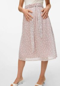 Una donna sta in piedi con le mani sui fianchi, indossando una gonna di un rosa chiaro con un motivo a foglie e tacchi a strappo bianchi.