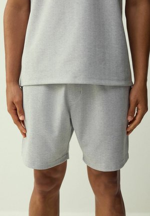 Hellgraue Shorts mit Fischgrätmuster und passendes Oberteil, getragen von einer Person, die mit entspannt an den Seiten hängenden Armen steht.