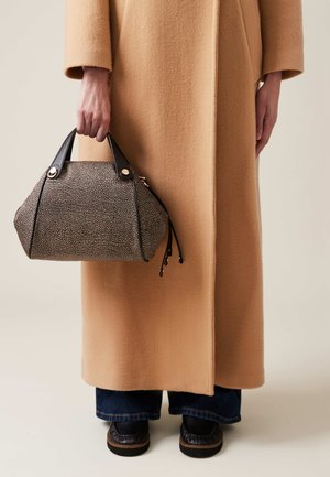 Texturiert schwarze und cremefarbene Handtasche mit gerundeter Form, schwarzen Ledergriffen und goldenen Akzenten, in der Hand gehalten, getragen mit einem hellbraunen Mantel.