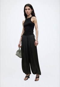 Γυναίκα με μακριά σκούρα μαλλιά, που φοράει μαύρο αμάνικο τοπ, φαρδιά μαύρη πλισέ παντελόνα, μαύρα ψηλοτάκουνα σανδάλια και κρατάει μια μικρή τσάντα με υφή.