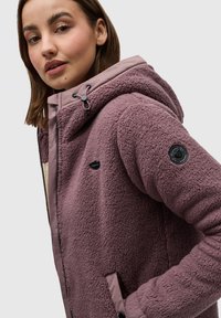 Veste en polaire de couleur mauve discret avec capuche, dotée d'une fermeture éclair, de poches latérales et d'un tissu texturé au toucher doux.