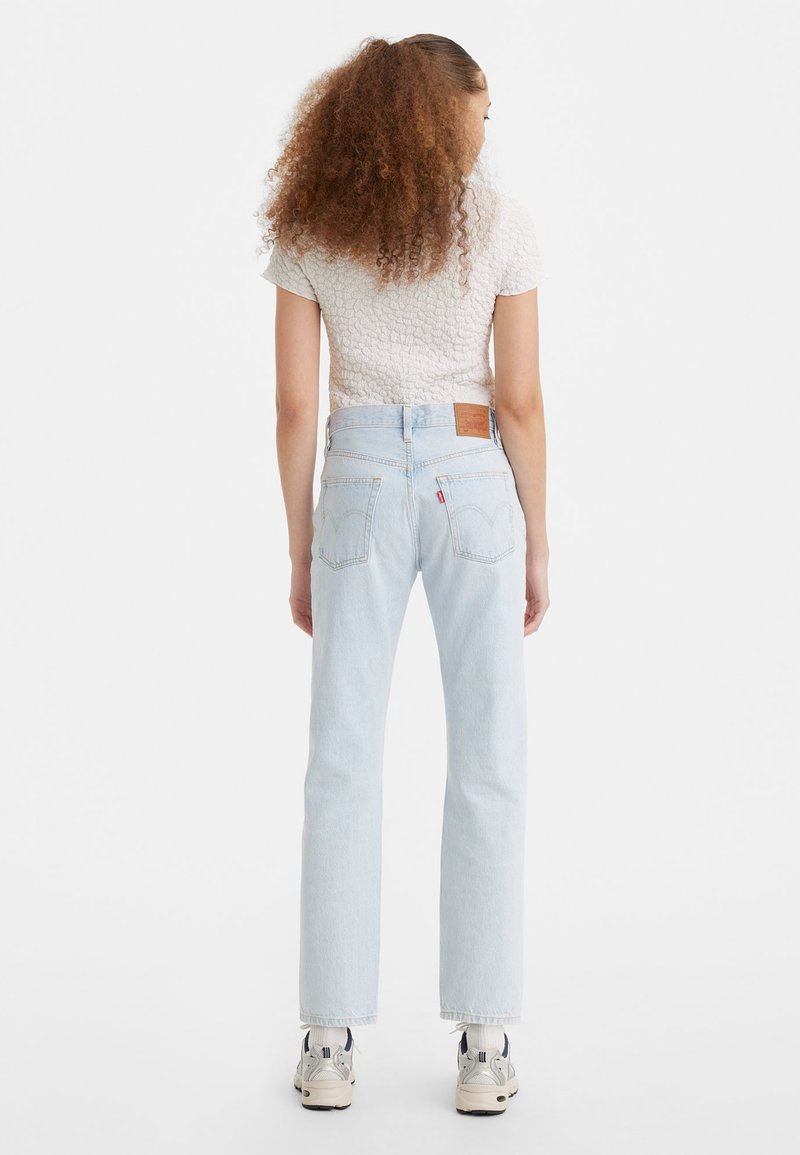 Femme aux cheveux bouclés de dos, portant un jean taille haute bleu clair, un haut blanc texturé à manches courtes, des chaussettes blanches et des baskets.