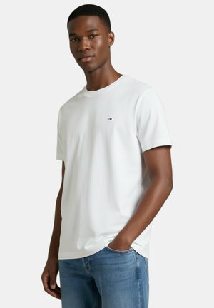 Joven vestido con una camiseta blanca de manga corta lisa con un pequeño logo en el pecho y jeans azules, de pie con una mano en el bolsillo.