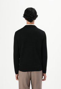 Man met kort krullend haar in een zwart lange mouwen trui en lichtbruine broek, staande met zijn rug naar de camera tegen een witte achtergrond.
