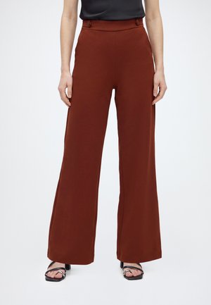 Femme portant un pantalon marron taille haute à jambes larges avec un détail de boutons à la taille et des sandales à talons noires ouvertes à l’avant.