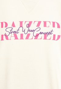 Prenda de color crema con atrevida letra en bloque rosa que dice "RAIZZED" y un "Concepto de Moda Callejera" en bordado morado.