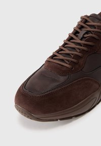 Brown suede shoe featuring a textured upper with mesh panels, round toe, and lace-up design. The sole has a subtle tread pattern.