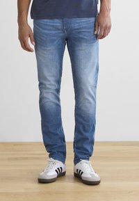 Person wearing light blue faded jeans and white sneakers with black stripes standing on a wooden floor.