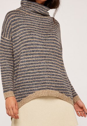 Femme portant un pull à col roulé ample rayé beige et gris avec un ourlet côtelé, assorti à une jupe couleur crème.