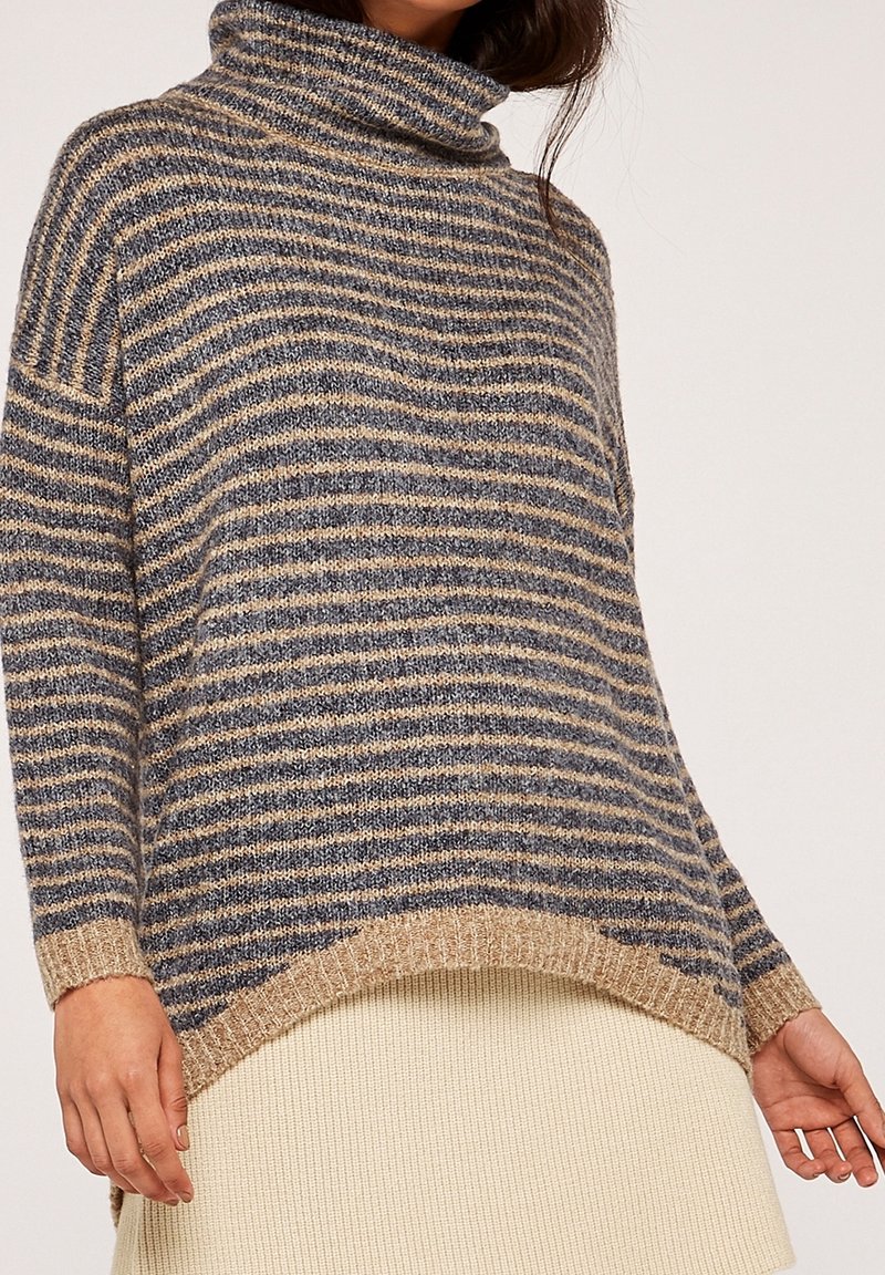Femme portant un pull à col roulé ample rayé beige et gris avec un ourlet côtelé, assorti à une jupe couleur crème.