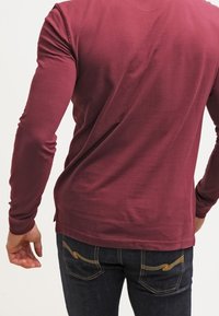 Bordeauxfarbendes Langarmhemd aus weichem Stoff, taillierte Passform, mit Rundhalsausschnitt und sichtbaren Nähdetails, kombiniert mit dunklen Jeans.