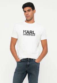 Vyras su trumpais garbanais plaukais, vilkintis baltą Karl Lagerfeld marškinėlį ir tamsiai mėlynas džinsus, stovi su rankomis kišenėse prieš baltą foną.