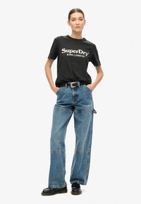 Camiseta de algodón negra con estampado "SuperDry SOHO.LONDON", combinada con jeans de mezclilla oversize de color azul claro que presentan paneles y un cinturón negro.