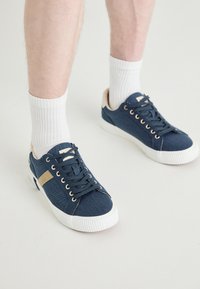 Navy blue sneakers with a denim texture, white rubber soles, gold eyelets, and a beige accent stripe. Worn with white ribbed socks.