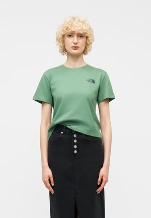 Femeie blondă purtând un tricou verde deschis The North Face și o fustă neagră cu talie înaltă și nasturi în față, stând în fața unui fundal simplu.