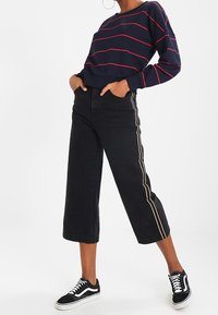 Sudadera de rayas navy con rayas rojas, pantalones cortos negros anchos con rayas doradas laterales y zapatillas negras con acentos blancos.