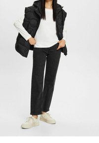 Czarna kamizelka puchowa na białej koszuli z długim rękawem, zestawiona z czarnymi dżinsami o szerokich nogawkach i beżowymi trampkami. Gładka faktura i swobodny styl.