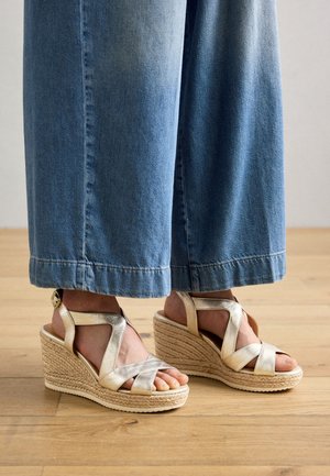Pies con sandalias de cuña doradas de tiras, con suela de plataforma trenzada, vistiendo vaqueros azules de pierna ancha sobre un suelo de madera.