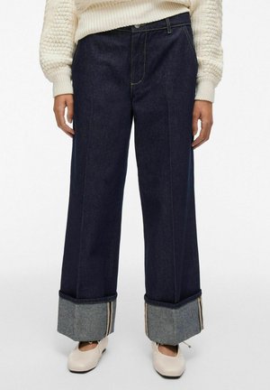 Persoon in donkere wijdlopende jeans met grote boorden, een crèmekleurige gebreide trui en crèmekleurige ballerina's.