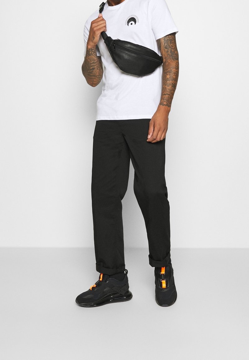 Riñonera de cuero negro llevada cruzada sobre el pecho, combinada con una camiseta blanca con un diseño gráfico, pantalones negros y zapatillas negras con acentos naranjas.