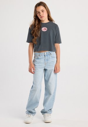 T-shirt gris coupé avec un logo rose circulaire, associé à un jean bleu clair à coupe décontractée et des baskets blanches. Design simple et texture du tissu.