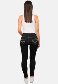 Zwarte skinny jeans met gouden stiksels, met een hoge taille en decoratieve achterzakken. Draagt een witte tanktop en witte sneakers.