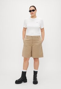 Weißes Kurzarmhemd mit kleinem Logo, beigefarbene knielange Shorts und schwarze Knöchelstiefel mit Plateausohlen. Getragen mit schwarzen Sonnenbrillen.