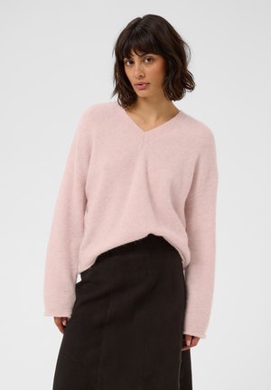Femme aux cheveux courts et foncés portant un pull rose clair à col en V et une jupe sombre taille haute, debout devant un fond uni.