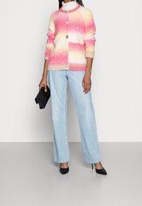 Cardigan lavorato a maglia con strisce sfumate rosa e gialle, colletto rotondo, chiusura frontale con bottoni e polsini a coste, abbinato a jeans a gamba larga azzurri e tacchi neri.