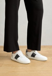 Sneakers bianchi con un design a zigzag nero sui lati, accenti grigi e superfici testurizzate, abbinati a pantaloni neri a gamba larga.