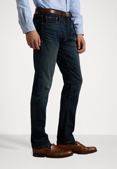 Pantalones vaqueros de denim azul oscuro con corte recto, que presentan un cinturón de cuero marrón, bolsillos laterales y un ligero desgastado en los muslos.