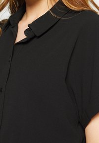 Blusa negra con cuello abotonado, mangas cortas y tejido texturizado. Presenta un diseño clásico con botones en la parte delantera y un acabado suave.