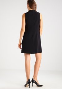 Czarna sukienka bez rękawów o dopasowanej sylwetce, z koralikowym kołnierzykiem, gładką fakturą i sięgająca powyżej kolan. Noszona z czarnymi szpilkami.