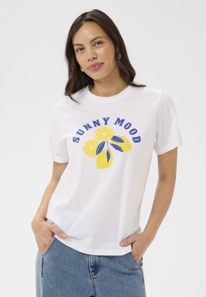 Femme portant un T-shirt blanc avec le texte "SUNNY MOOD" et un graphisme de citron jaune, associée à un jean bleu, posant les mains dans les poches.