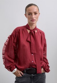 Blusa roja con volantes, con un lazo en el cuello y cierre de botones. Presenta mangas fluidas y translúcidas, y una tela texturada. Combinada con un cinturón negro.