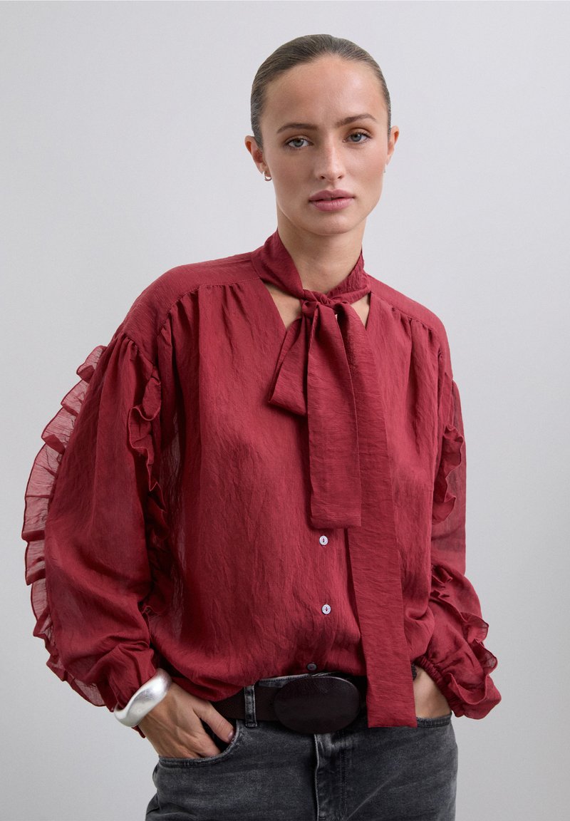 Blusa roja con volantes, con un lazo en el cuello y cierre de botones. Presenta mangas fluidas y translúcidas, y una tela texturada. Combinada con un cinturón negro.